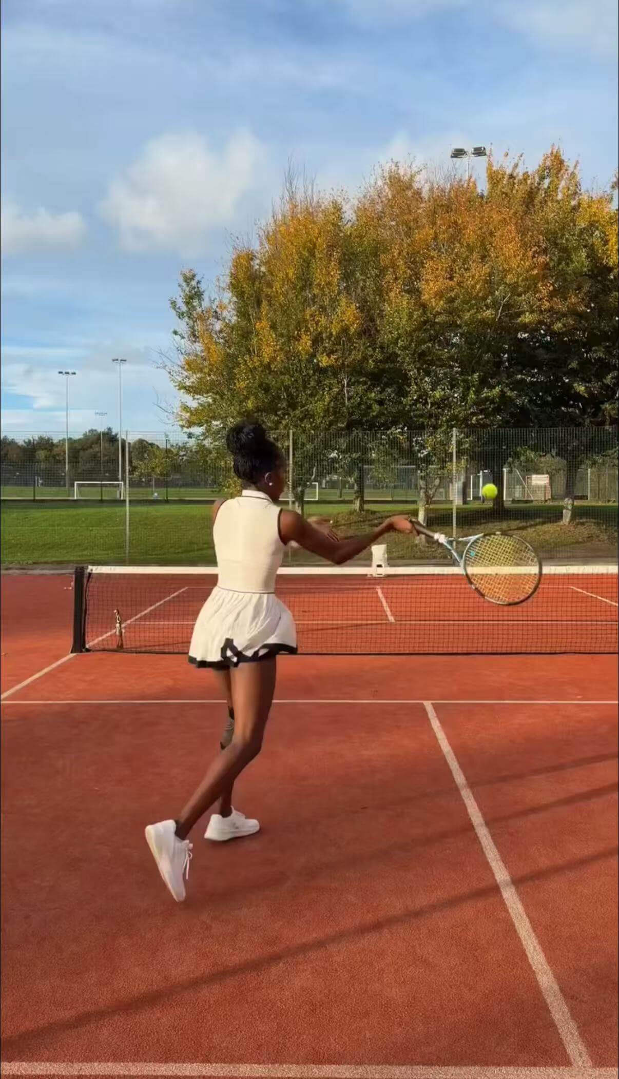 thereal.hms playing tennis on a clay court with trees and blue sky in the background