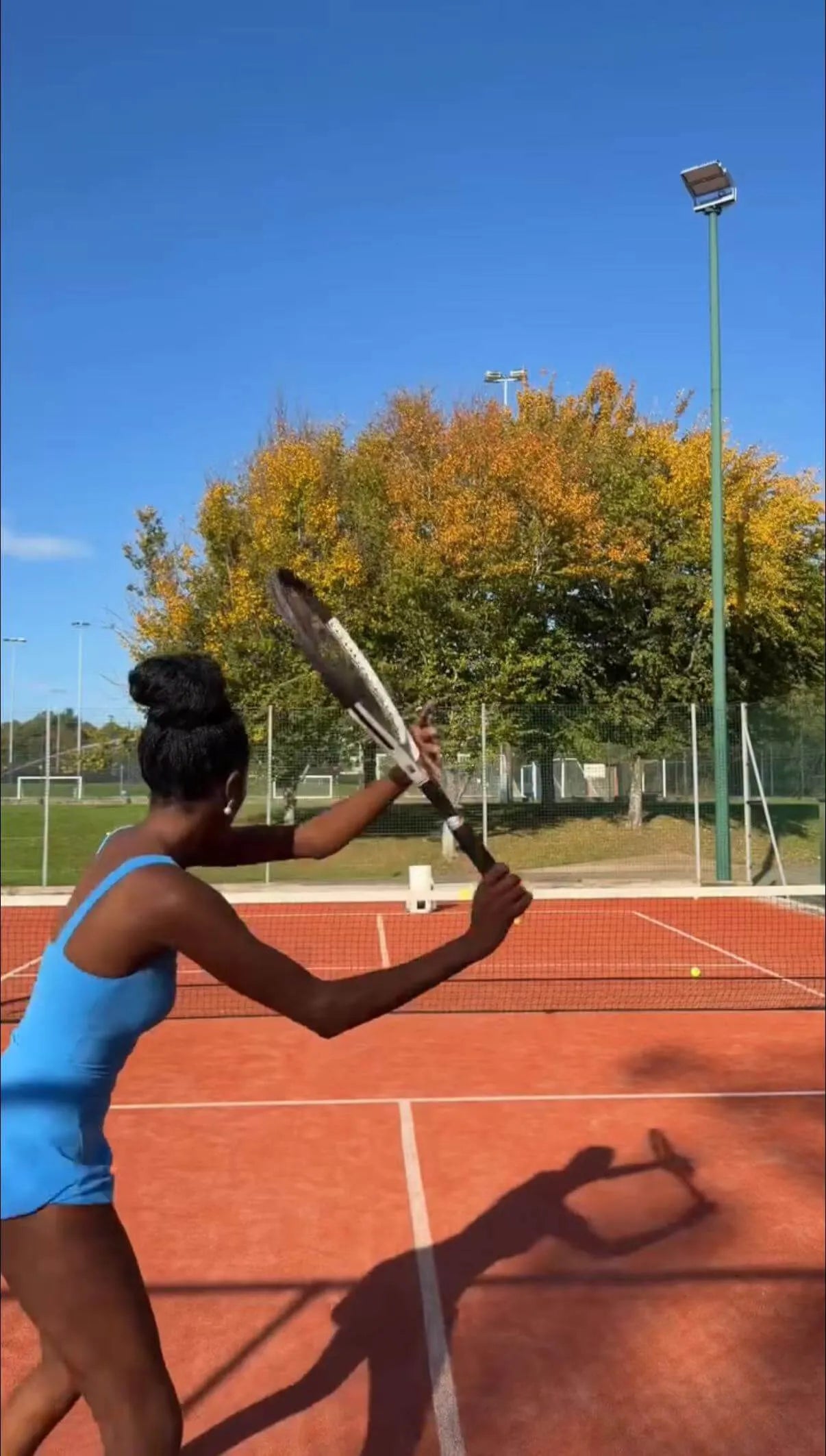 thereal.hms playing tennis on a clay court with trees and blue sky in the background